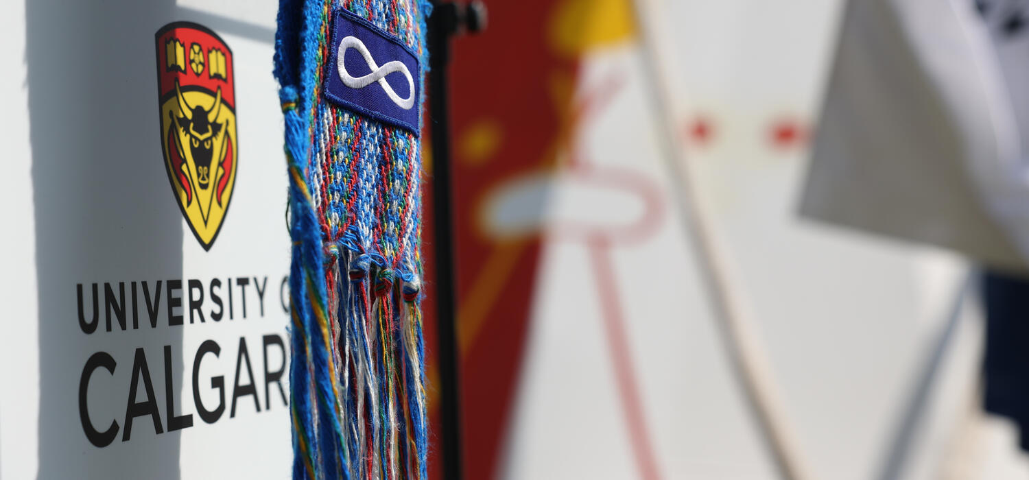 A blue Ceinture Fl茅ch茅e (M茅tis arrow sash) featuring the M茅tis flag hangs in front of the UCalgary logo. In the background a tepee is visible.