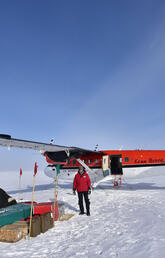 A man in a red jacket stands in front of a red plane with luggage stacked next to him on a snowy landscape